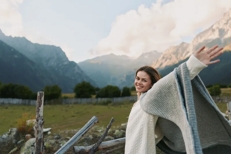 A smiling woman in a cozy knitted sweater enjoys fresh air outdoors with arms outstretched against a mountain landscape. Concept of freedom and lifestyle.