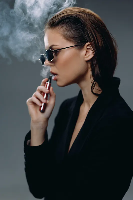 Stylish woman vaping with sunglasses and black jacket exhaling smoke in dark background