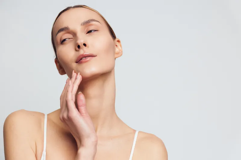 Young woman with clear skin gently touching her face with closed eyes on a white background. Concept of skincare, beauty, wellness, and natural health care.