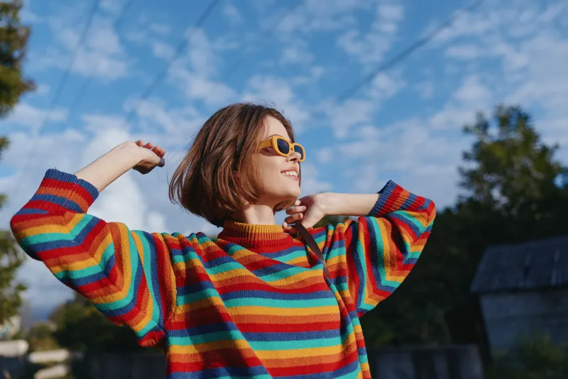 woman in colorful striped sweater wearing sunglasses smiles outdoors in a sunny portrait, short hair and relaxed pose against blue sky for fashion, lifestyle and joy