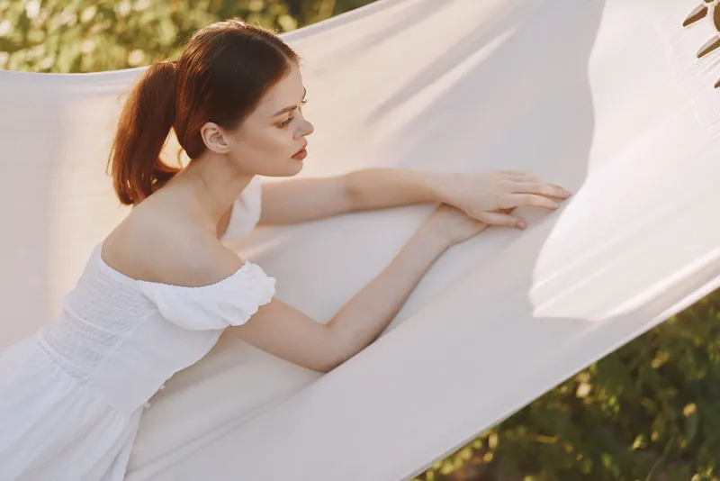 A serene young woman in a white dress relaxing on a hammock, embracing nature s tranquility with soft sunlight filtering through leaves, evoking feelings of peace and calm