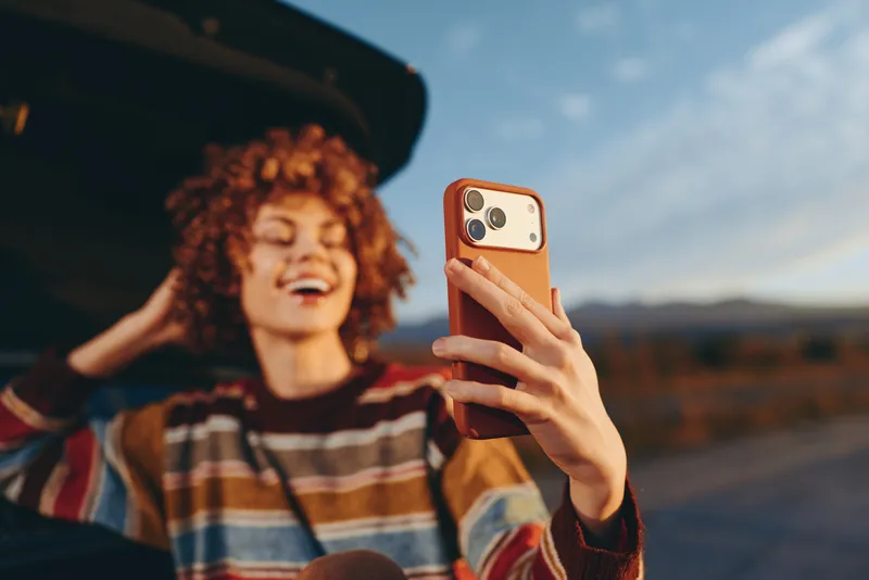 Woman lifestyle smartphone selfie, outdoor sunset, travel leisure, curly hair smiling, closeup hand holding phone screen, technology, blurred mountains landscape behind, evening sunlight