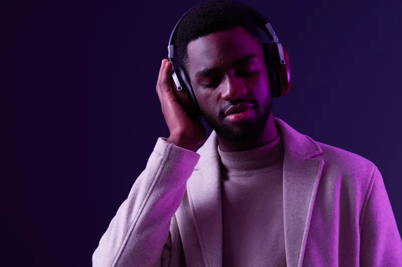 A young Black man enjoying music with headphones, dressed in a stylish beige coat and light sweater, set against a vibrant purple background, conveying relaxation and contentment