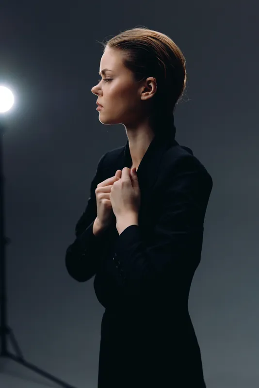 Profile of a young woman in a black jacket with a serious expression on a dark background with soft studio lighting.