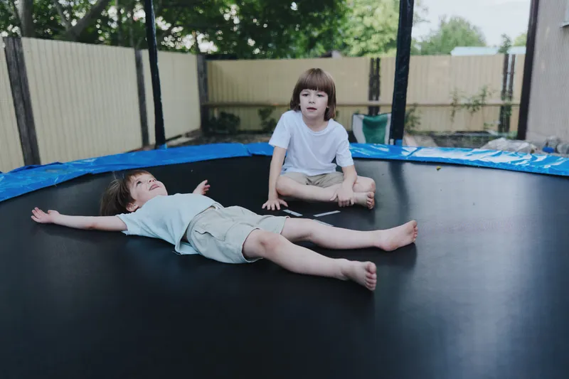Two happy children playing on a trampoline, one lying down while the other sits nearby, showcasing joy and laughter in a sunny outdoor setting.