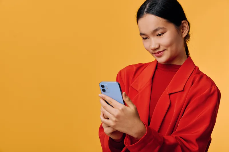 Young woman in red jacket using smartphone against yellow background, smiling and texting message with interest and focus on digital device in casual modern style portrait photo.