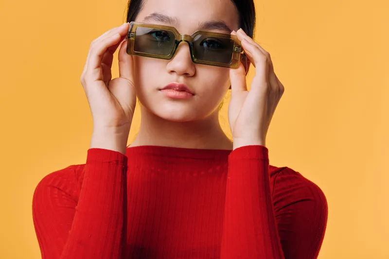 Young woman in red sweater adjusting sunglasses with both hands against yellow background. Fashion portrait, casual style, confident female model with modern eyewear studio shot.