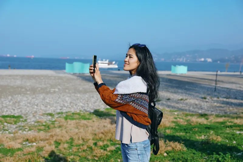 Woman selfie beach outdoors, holding a phone against a coastal backdrop with sea, sand, and blue sky, capturing leisure travel vacation vibes in a candid moment on a sunny shore