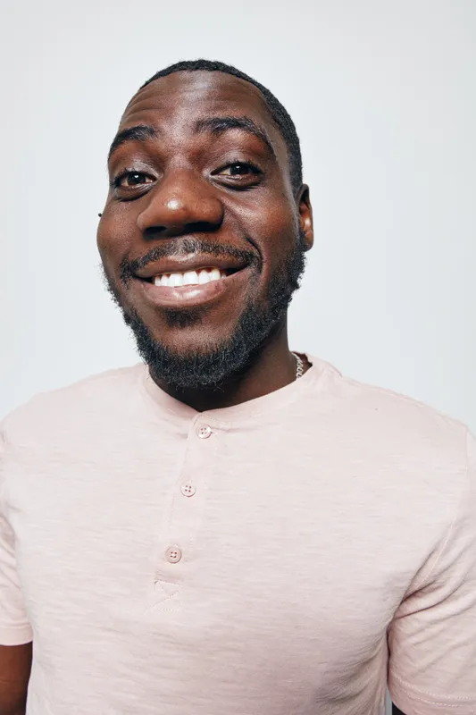 African American man with dark skin smiling and wearing light pink shirt standing against white background. Portrait of happy male with beard and short hair looking at camera.