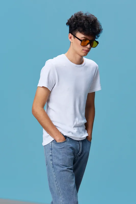 young man wearing sunglasses and casual white t-shirt standing with hands in pockets against blue background, relaxed and stylish urban fashion portrait