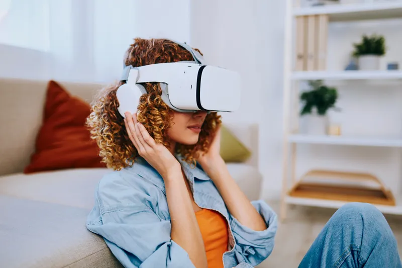 Улыбающаяся женщина наслаждается игрой в виртуальной реальности дома в футуристических очках VR на диване