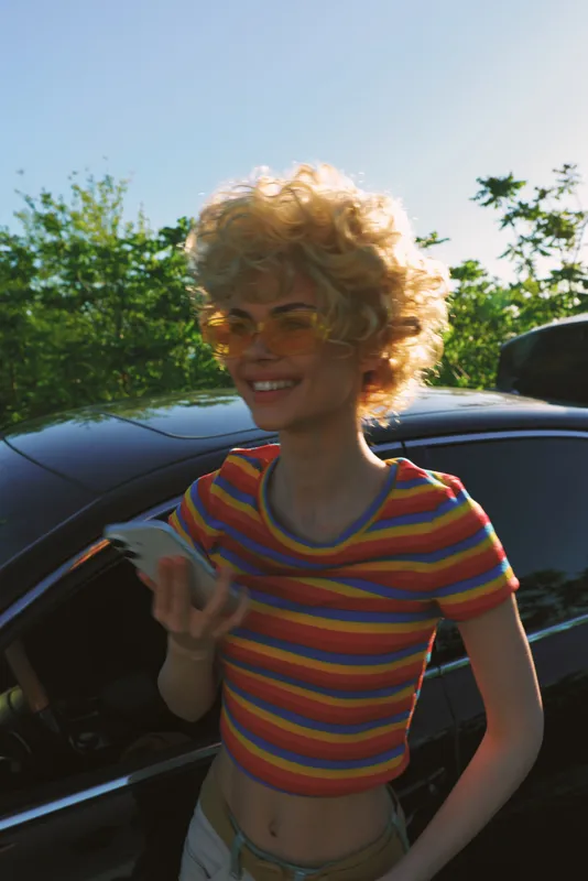 Smiling woman with curly blonde hair in a striped crop top stands beside a car in a sunlit outdoor scene, conveying casual lifestyle, confidence, and retro warmth.