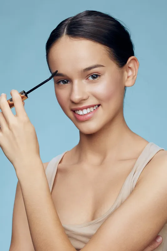 Smiling young woman applying brow gel with a blue background, showcasing beauty and confidence while promoting self care and makeup techniques