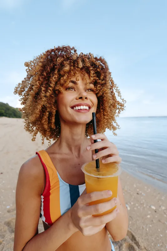 Curly hair woman smiling on a beach, holding a cold drink in a glass bottle, sunny sky and sea behind, summer vacation mood, relaxed summer refreshment, natural outdoor light