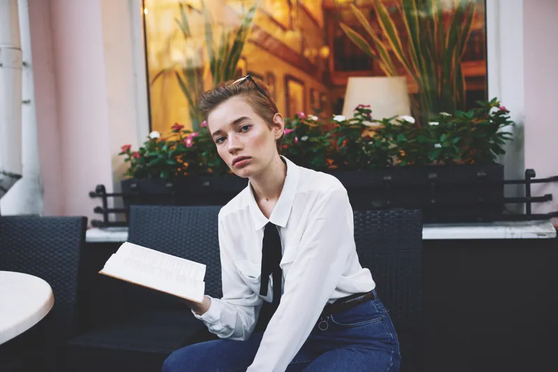 Красивая молодая женщина с короткими волосами в очках держит в руках книгу, сидя за столиком в кафе