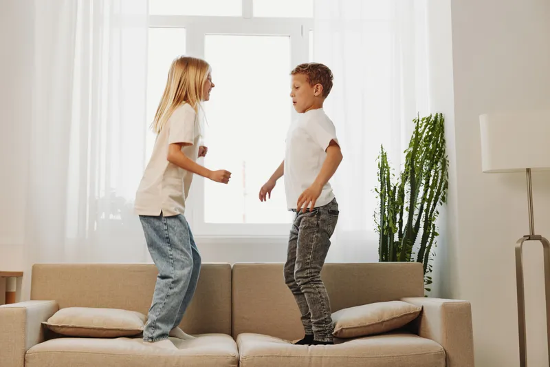 Two happy children jumping on beige sofa in bright living room near window with green plant. Concept of childhood fun and energetic play indoors.