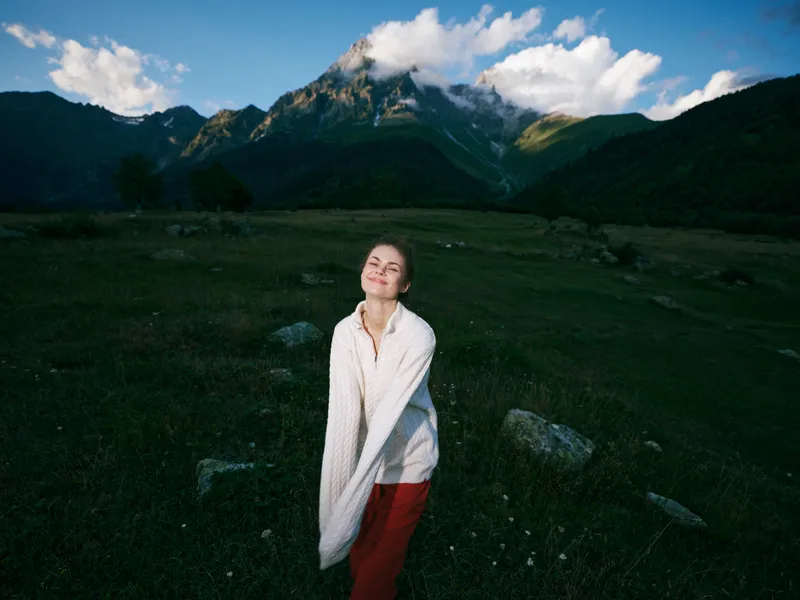 A woman standing in a grassy meadow with a dramatic mountain landscape, wrapped in a light blanket, enjoying fresh air and distant peaks in a peaceful outdoor moment