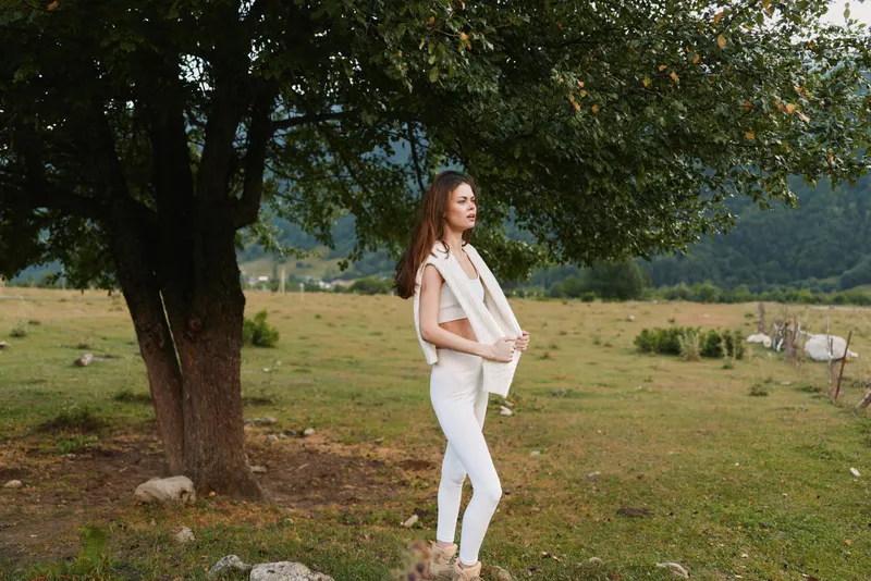 Young woman with long curly hair stands thoughtfully under a large tree in a green meadow wearing white pants and a sleeveless top with a light jacket over her shoulders.