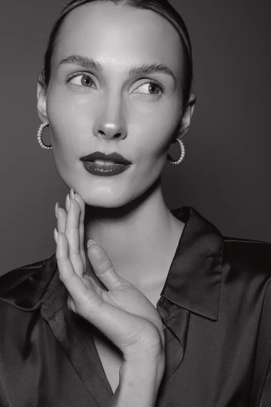 Portrait of a light skinned white woman with slicked hair and glossy lips wearing hoop earrings and a satin blouse. Elegant female model poses in black and white studio photo.
