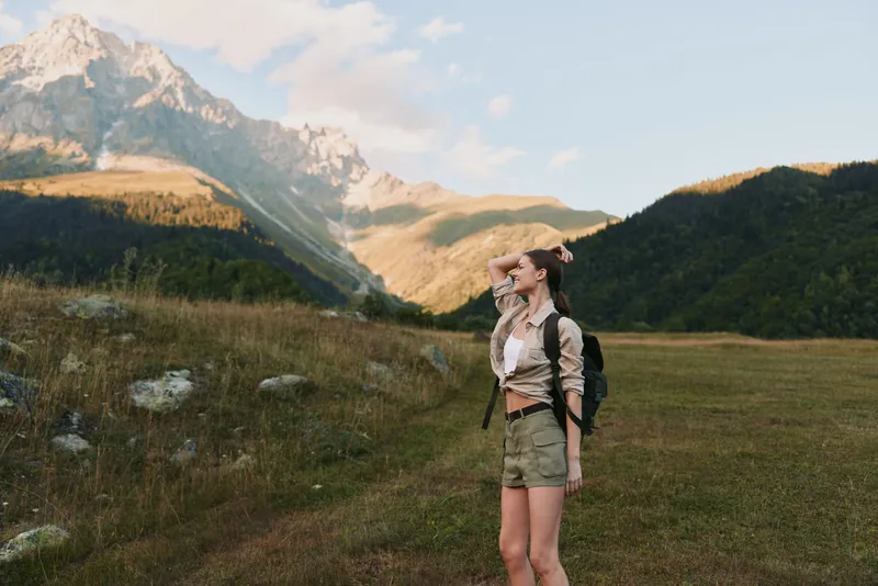 A young woman with a backpack stands in a sunlit meadow, looking toward distant mountains, enjoying a moment of outdoor adventure and freedom.