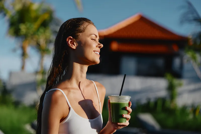 Cheerful woman outdoors, smiling with eyes closed, sunlit moment, casual summer vibe, fresh beverage in hand, breezy hair, warm lighting, lush garden backdrop