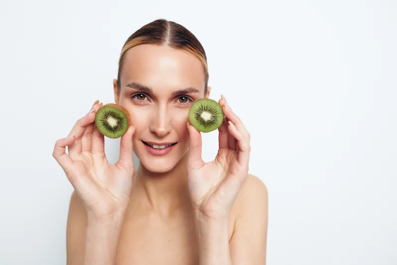 Woman holding kiwi fruit slices near cheeks, smiling in a beauty skincare portrait, fresh wellness mood, natural skin, bright daylight, clean white background, healthy diet idea, fruit beauty