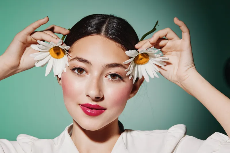 Young woman with clear skin holds two white daisies by her face, showcasing glossy pink lips and smooth makeup on a pastel green background. Concept for beauty, fashion, and creativity.