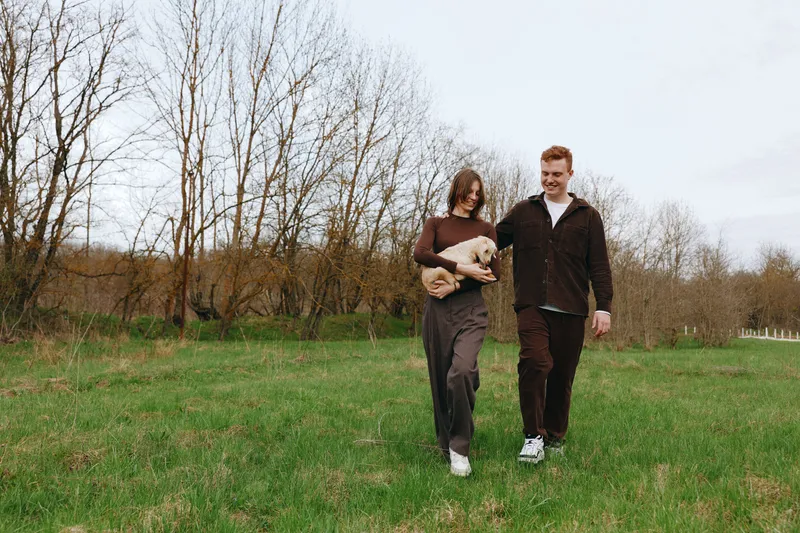 A couple walks outdoors in a grassy field with a gentle goat kid, sharing a calm moment. A lifestyle image showing companionship, nature, and rural living with an animal in the frame.