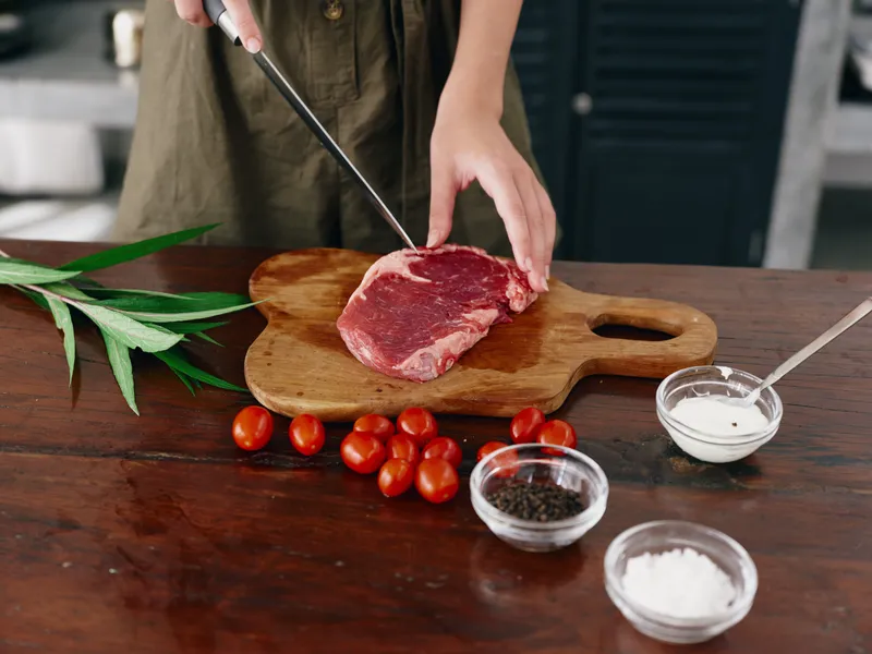 Женщина с ножом в руке нарезает мясо для стейка для жарки на кухне, на столе соль, перец и другие специи, красные помидоры черри и зелень, приготовление ужина