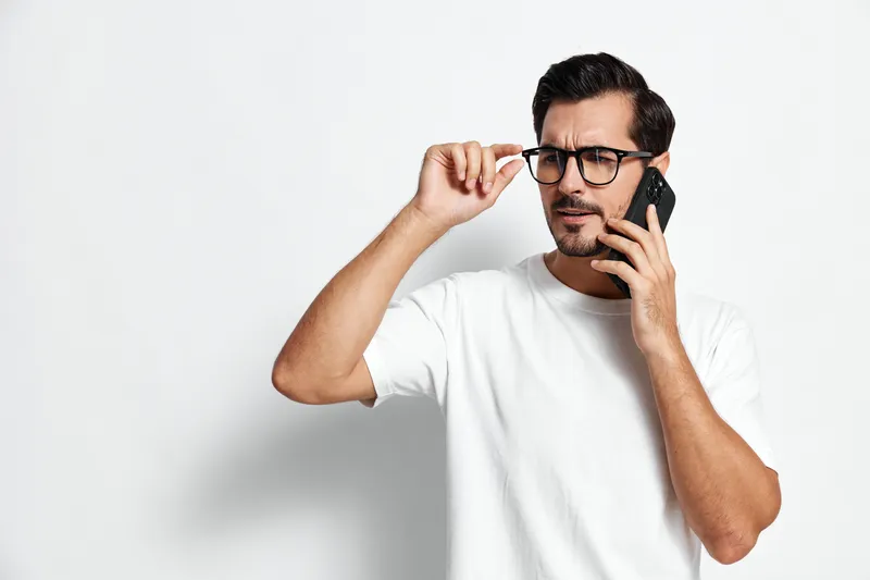 Thoughtful man wearing glasses holding mobile phone talking while standing against plain solid white background, looking serious. People communication concept.