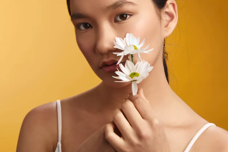 asian woman smiles subtly as she holds white daisies near her lips in a modern studio, warm yellow backdrop, soft lighting, capturing gentle beauty and a calm, natural vibe.
