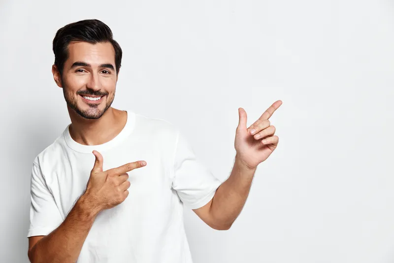 Smiling happy young man in a white t-shirt pointing with fingers at an empty space, looking at the camera with a cheerful expression, isolated on a plain background.
