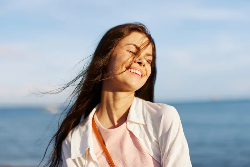 Девушка с закрытыми глазами на солнце на фоне океана улыбается, показывая зубы, развевающиеся волосы, загорелая кожа, отдыхает, путешествует к океану