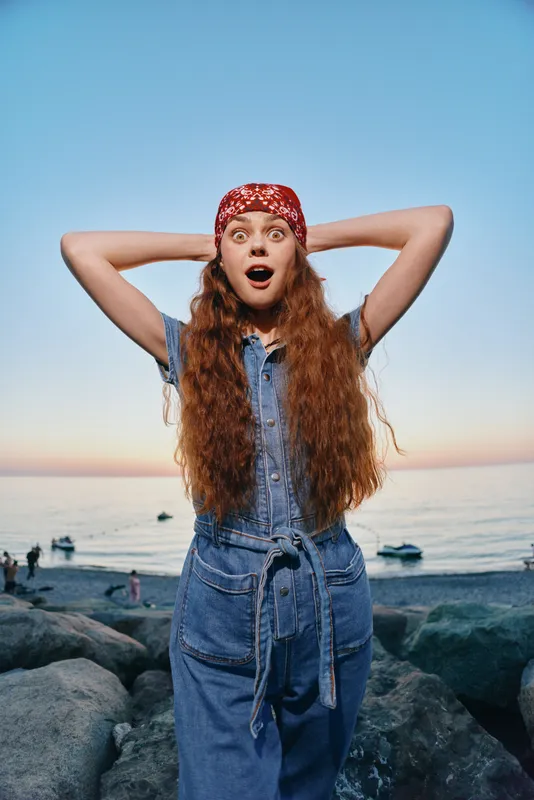 surprised woman with long curly hair wearing denim outfit and red bandana standing on rocks by the sea at sunset with clear sky
