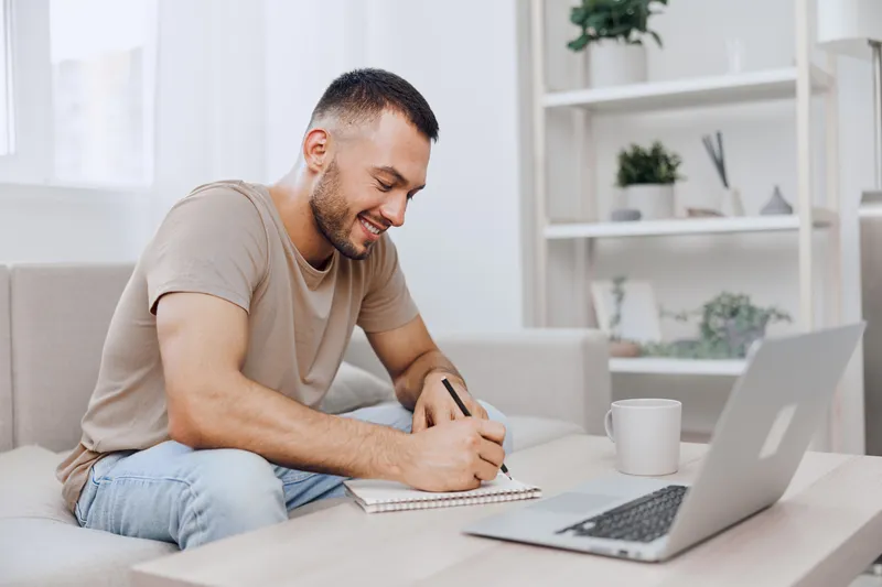 Smiling man writing in a notebook, showcasing modern workspace aesthetics with natural light and minimalism, perfect for remote work inspiration.