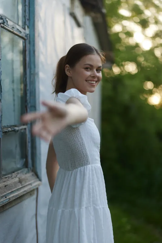 Young woman in a white dress reaching out with a smile, surrounded by greenery, conveying a sense of joy and openness during a sunny day