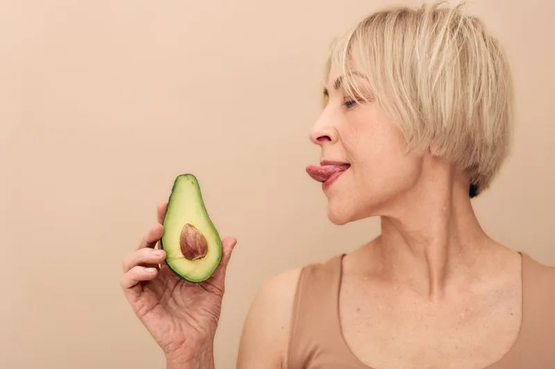 Mature woman with short blonde hair holding half an avocado close to her face, playfully sticking out her tongue. Concept of healthy eating, nutrition, and natural lifestyle.