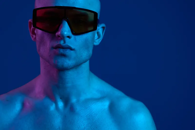 A striking portrait of a man wearing stylish sunglasses against a vibrant blue background. The contrast of colors highlights his strong features and bold expression.