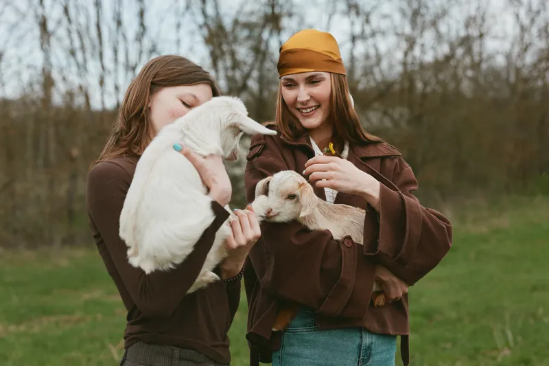 Cheerful women outdoors, laughing with goat kid, gentle feeding moment, natural colors, soft breeze, casual clothes, sunny day, expansive meadow background, candid lifestyle scene, warm animal