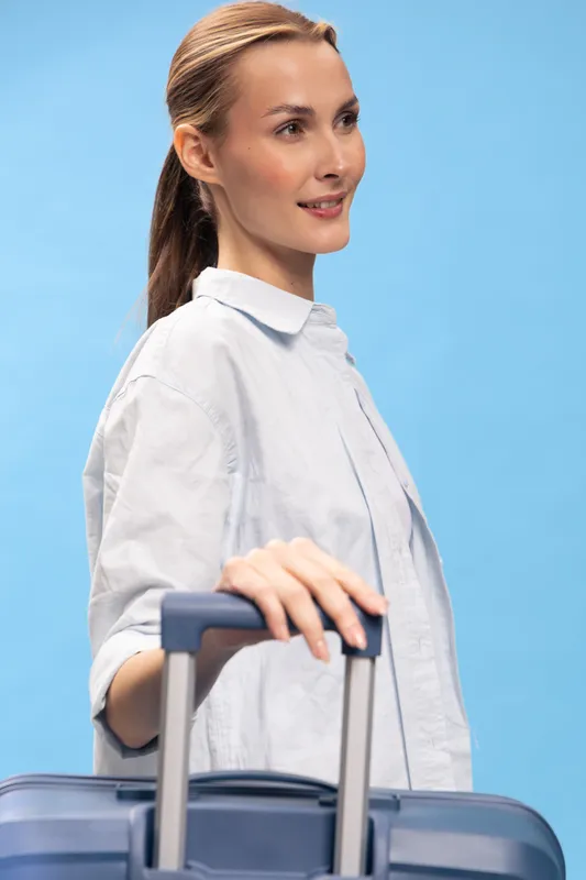 Young woman in white shirt holding a blue suitcase handle against a blue background. Concept of travel, vacation, and summer trip with luggage and casual style in studio lighting.