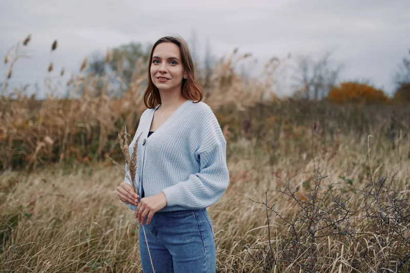 A calm, genuine portrait of a woman standing in a windswept field, wearing a soft knit cardigan and jeans, conveying trust, authenticity through a natural pose and quiet confidence.
