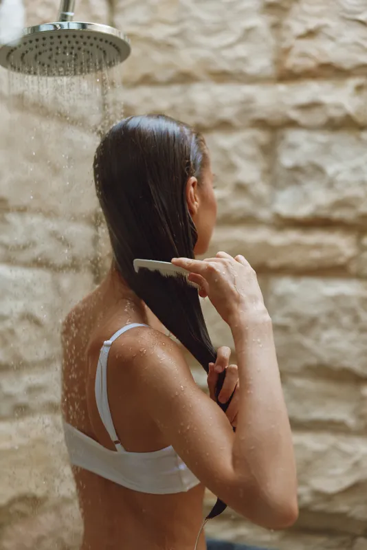 woman combing wet hair under shower with stone wall background, intimate moment, personal care, beauty routine, natural light, calm atmosphere