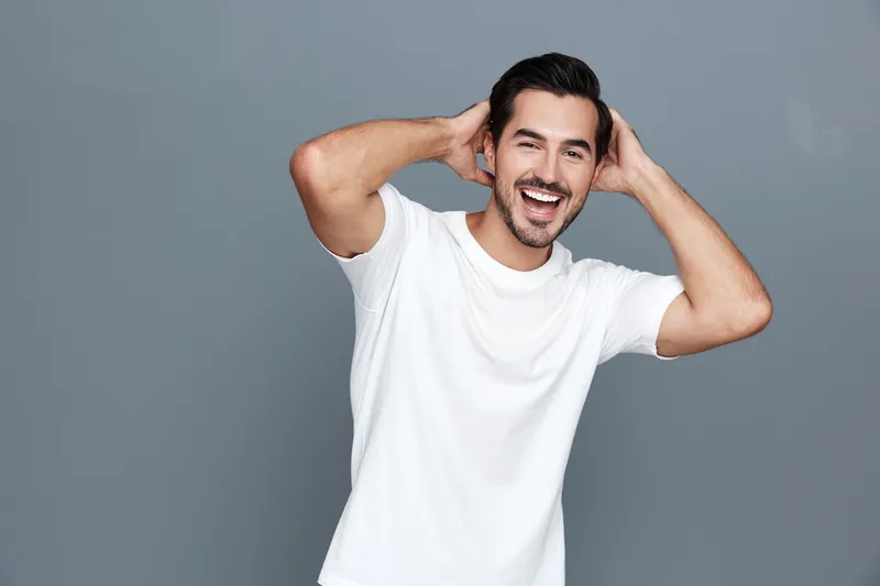 Full body cheerful happy young man smiling with hands behind head, wearing a casual white t-shirt, posing against a solid gray background. Lifestyle concept.