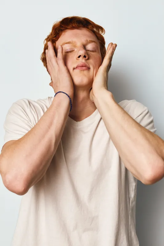 Young man with red hair in white shirt touching face with eyes closed showing relaxation and calm expression on light background. Skin care, wellness and peaceful moment.