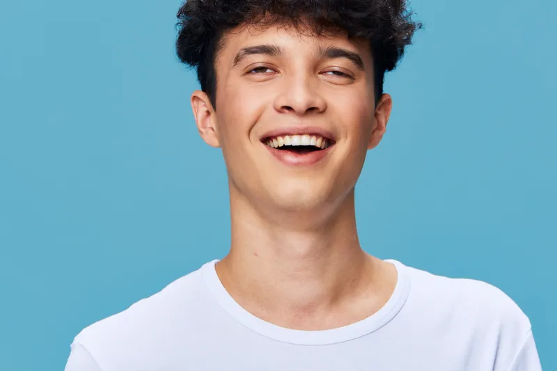 smiling young man in white t-shirt with curly hair laughing against blue background, joyful expression and casual style portrait, bright and clean setup