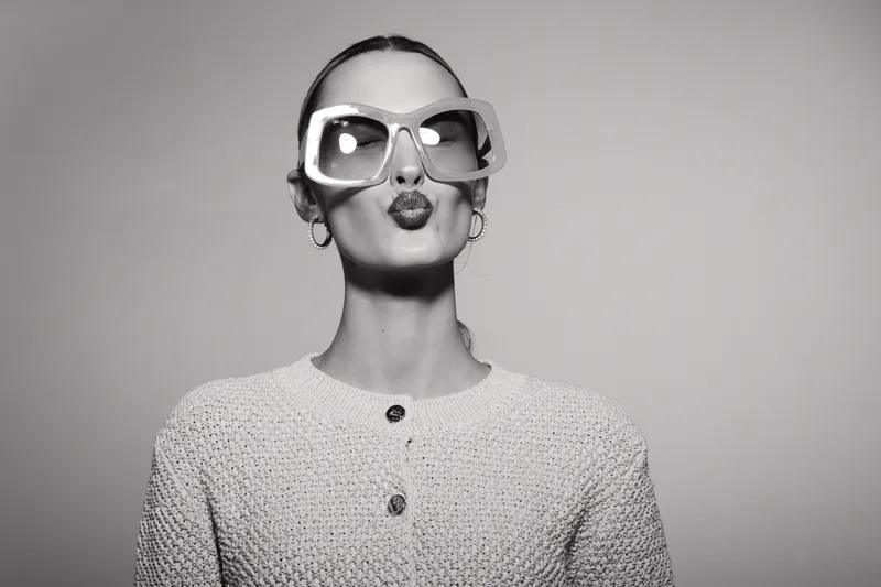 Stylish young Caucasian woman with pale skin and slicked back hair wearing large square sunglasses and a knitted cardigan, making a playful kiss gesture on a plain background.