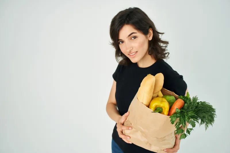 Женщина держит в руках бумажный пакет с продуктами, доставляя овощи и здоровую пищу