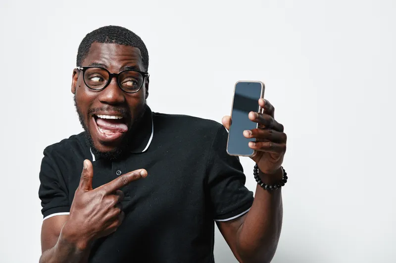 African American man with dark skin and glasses excitedly pointing at smartphone screen, showing technology use and communication with happy expression on white background.