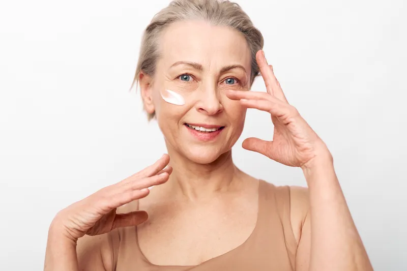 Mature woman with light skin applies face cream on cheek, smiling gently. Close-up portrait on white background. Concept of skincare, aging beauty, and self-care routine.