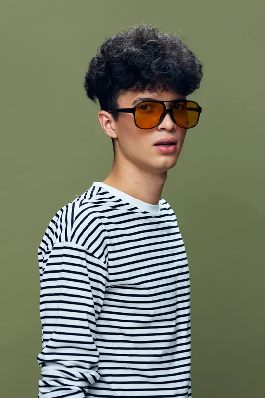 young man wearing amber sunglasses and striped shirt with curly black hair standing against simple green background, looking sideways in casual style and natural light